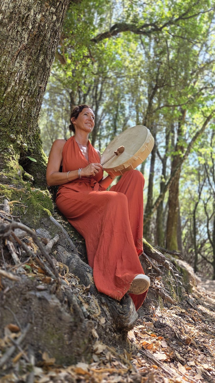 Céline Dusseval assise contre un arbre, son tambour chamanique à la main
